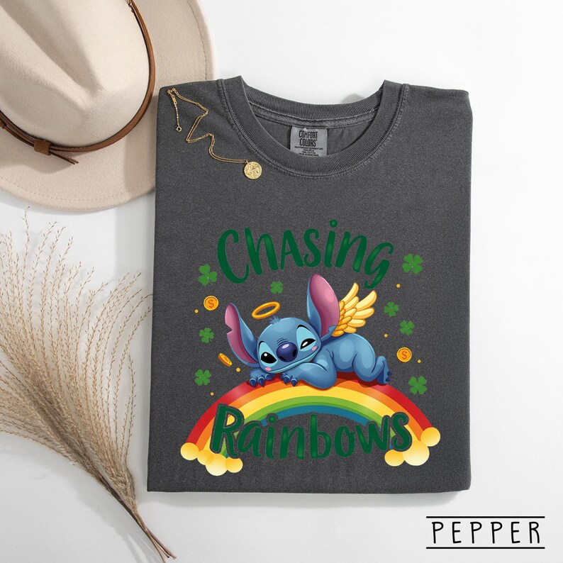 May include: Dark grey t-shirt with a cartoon character with angel wings and a halo, resting on a rainbow. The shirt has the words "Chasing Rainbows" in green and yellow. Gold coins and shamrocks are also featured.
