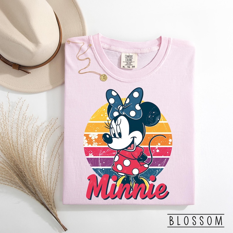 May include: A light pink Comfort Colors t-shirt featuring a Minnie Mouse graphic. The graphic includes Minnie in a red polka dot dress and a blue polka dot bow, set against a retro sunset design. The word "Minnie" is written in red script.