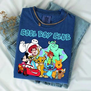 Może przedstawiać: Niebieski t-shirt z kr&oacute;tkim rękawem z napisem COOL BOY CLUB jasnoniebieskimi literami. Grafika przedstawia postacie Disneya: Baymaxa, Chudego, Myszkę Miki, Zygzaka McQueena, Simbę, Nicka Wilde'a, Sulleya, Sticha, Kubusia Puchatka i Remy'ego.