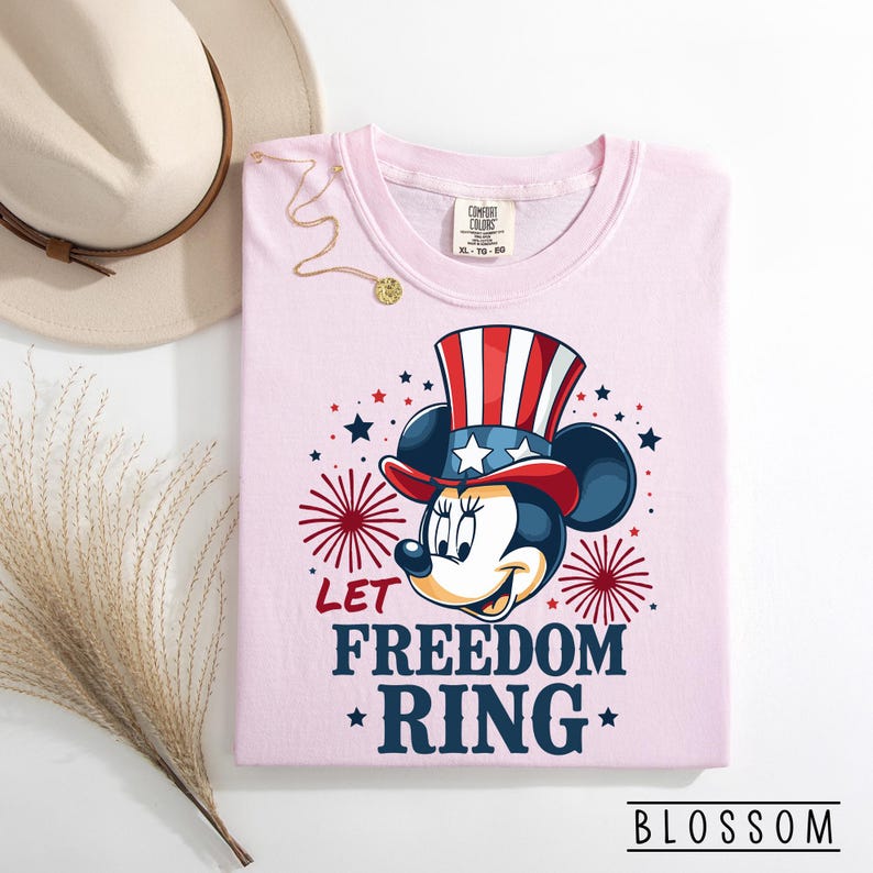 May include: A light pink t-shirt featuring a patriotic Minnie Mouse design. Minnie Mouse is wearing a red, white, and blue Uncle Sam hat. The text "LET FREEDOM RING" is below the graphic, with red and blue stars and fireworks.