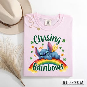 May include: A pale pink t-shirt with the words "Chasing Rainbows" and a cartoon character lying on a rainbow. The shirt also features shamrocks and gold coins. The brand name "Blossom" is at the bottom.