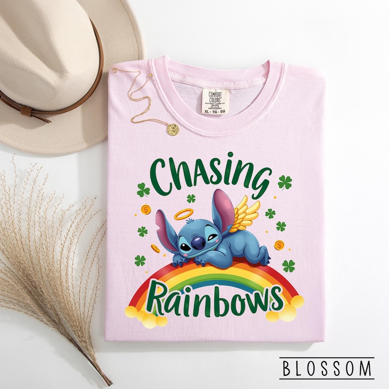 May include: A pale pink t-shirt with the words "Chasing Rainbows" and a cartoon character lying on a rainbow. The shirt also features shamrocks and gold coins. The brand name "Blossom" is at the bottom.