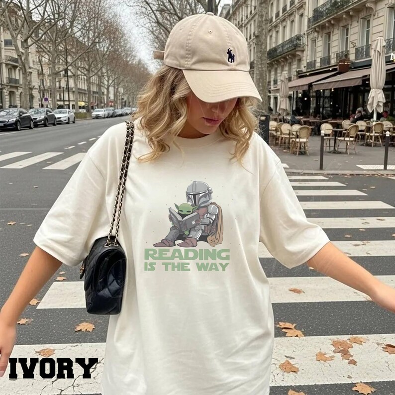 Puede incluir: Camiseta de color marfil con un gr&aacute;fico de The Mandalorian y Grogu leyendo un libro, con el texto verde "READING IS THE WAY". La persona lleva una gorra de b&eacute;isbol beige con un logotipo negro y un bolso negro.