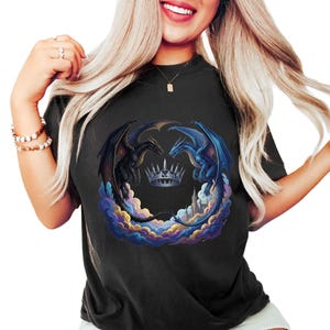 May include: Black t-shirt featuring a fantasy design with two dragons facing each other, a crown, and a cityscape. One dragon is black, the other is blue. The design is surrounded by colorful clouds.