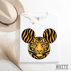 May include: White t-shirt featuring a tiger face design with Mickey Mouse ears. The tiger design is yellow and black. The shirt has a crew neck and the word "WHITE" is printed at the bottom.