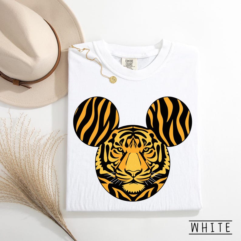May include: White t-shirt featuring a tiger face design with Mickey Mouse ears. The tiger design is yellow and black. The shirt has a crew neck and the word "WHITE" is printed at the bottom.