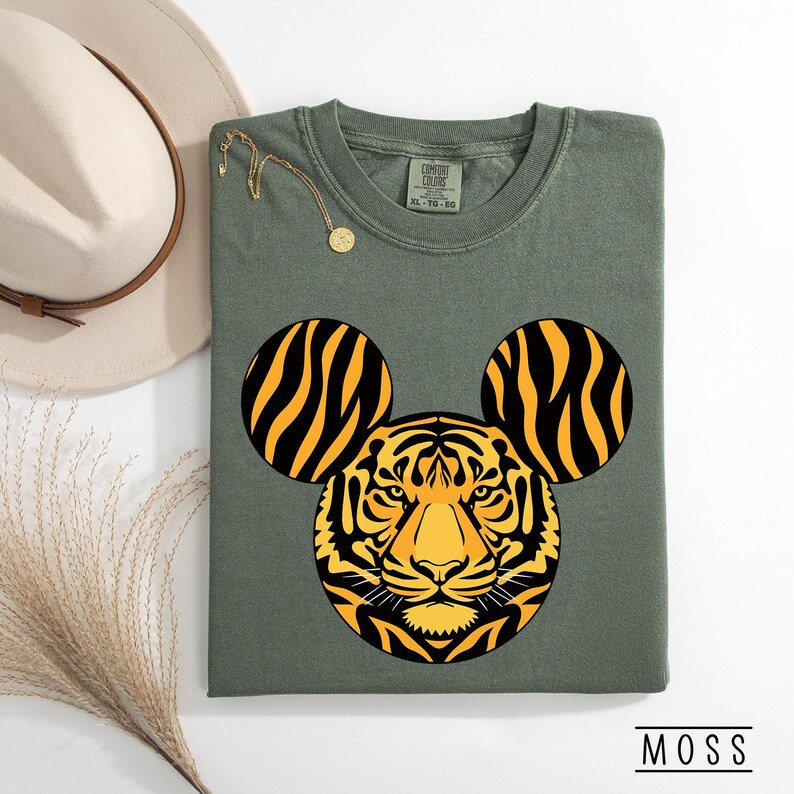 May include: Olive green t-shirt featuring a tiger design with Mickey Mouse ears. The tiger's face is yellow and black, with black stripes on the ears. The shirt has a crew neck and short sleeves. The text "COMFORT COLORS" is visible on the tag.