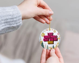May include: A round Christmas ornament with the text "LAST CHRISTMAS I GAVE YOU MY RAGE" in pink and white. The ornament has gold and green Christmas tree and ornament designs. It is held by a gold string.