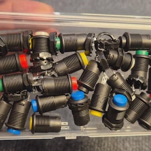 May include: A clear plastic container filled with black push button switches. The buttons have various colored tops, including red, green, yellow, and blue. The buttons are cylindrical with a threaded body.