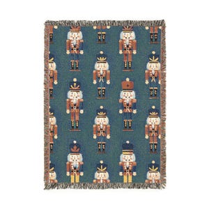 May include: A woven throw blanket with a repeating pattern of nutcracker figures in red, blue, and white on a teal background. The blanket has a fringed edge in shades of brown and tan. A festive holiday home decor item.