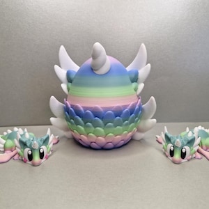 May include: A set of whimsical fantasy creature figurines. The central figure is egg-shaped with a rainbow gradient and white wings. Two smaller dragon-like figures with pastel colours and wings are positioned on either side, creating a playful scene.