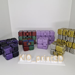 Puede incluir: Una colecci&oacute;n de coloridos cubos fidget en varios colores, incluyendo morado, dorado, plateado, rojo y verde. Los cubos est&aacute;n dispuestos sobre una superficie blanca, con el texto "KIO_print3D" en dorado.