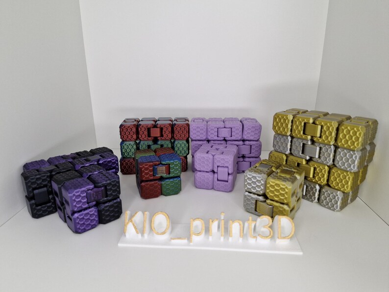 Puede incluir: Una colecci&oacute;n de coloridos cubos fidget en varios colores, incluyendo morado, dorado, plateado, rojo y verde. Los cubos est&aacute;n dispuestos sobre una superficie blanca, con el texto "KIO_print3D" en dorado.