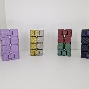 Puede incluir: Se muestran cuatro juguetes fidget de colores. Los juguetes est&aacute;n hechos de bloques conectados en p&uacute;rpura, dorado, rojo y verde, y negro y p&uacute;rpura. Cada juguete est&aacute; de pie, mostrando su esquema de color y dise&ntilde;o &uacute;nicos.