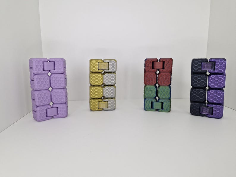 Puede incluir: Se muestran cuatro juguetes fidget de colores. Los juguetes est&aacute;n hechos de bloques conectados en p&uacute;rpura, dorado, rojo y verde, y negro y p&uacute;rpura. Cada juguete est&aacute; de pie, mostrando su esquema de color y dise&ntilde;o &uacute;nicos.