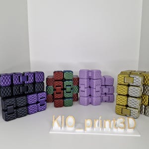 Puede incluir: Una colecci&oacute;n de coloridos juguetes fidget en varios colores, incluyendo morado, negro, rojo, verde y dorado. Los juguetes est&aacute;n dispuestos en pilas y est&aacute;n hechos de cubos entrelazados. El texto "KIO_print3D" se muestra en una base blanca.