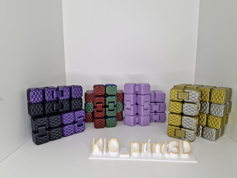 Puede incluir: Una colecci&oacute;n de coloridos juguetes fidget en varios colores, incluyendo morado, negro, rojo, verde y dorado. Los juguetes est&aacute;n dispuestos en pilas y est&aacute;n hechos de cubos entrelazados. El texto "KIO_print3D" se muestra en una base blanca.