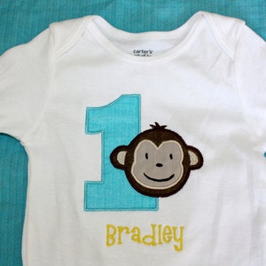 May include: White long-sleeve onesie with a blue number one and a brown monkey face. The onesie has the name "Bradley" in yellow letters.