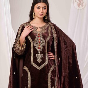 May include: A brown velvet salwar kameez with intricate gold embroidery and red bead accents. The ensemble includes a matching shawl. The outfit features a keyhole neckline and ornate detailing, showcasing a traditional design.