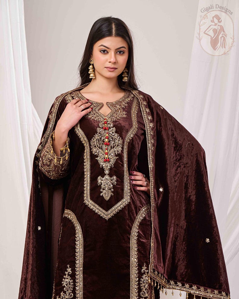 May include: A brown velvet salwar kameez with intricate gold embroidery and red bead accents. The ensemble includes a matching shawl. The outfit features a keyhole neckline and ornate detailing, showcasing a traditional design.