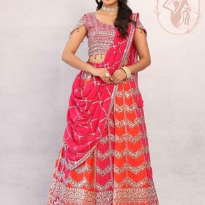 May include: A vibrant pink and orange lehenga with intricate silver embroidery. The outfit features a matching pink blouse with embellished sleeves and a draped dupatta. The design includes a detailed skirt with a chevron pattern.