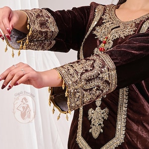 May include: A dark brown velvet garment with intricate gold embroidery and embellishments. The sleeves and neckline are adorned with detailed patterns and hanging gold beads. The garment features a decorative design and is likely part of a traditional outfit.