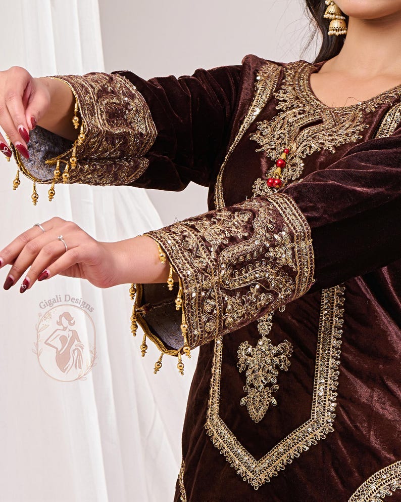 May include: A dark brown velvet garment with intricate gold embroidery and embellishments. The sleeves and neckline are adorned with detailed patterns and hanging gold beads. The garment features a decorative design and is likely part of a traditional outfit.