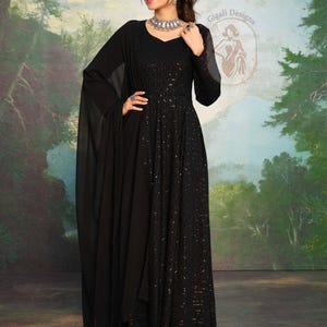 May include: A full-length black dress with long sleeves and a flowing skirt. The dress has detailed sequin embellishments and a matching shawl. The model is wearing silver jewellery. The background is a painted landscape with trees.