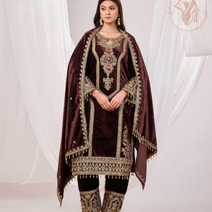 May include: A dark brown velvet salwar kameez ensemble with intricate gold embroidery. The outfit includes a long tunic, matching pants, and a draped shawl. The embroidery features floral and geometric patterns, with red and gold accents.