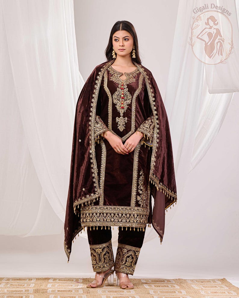 May include: A dark brown velvet salwar kameez ensemble with intricate gold embroidery. The outfit includes a long tunic, matching pants, and a draped shawl. The embroidery features floral and geometric patterns, with red and gold accents.