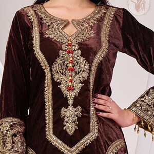 May include: A brown velvet tunic with intricate gold embroidery around the neckline, bodice, and cuffs. The design includes a keyhole neckline and is embellished with red beads. The sleeves are long, and the garment has a luxurious appearance.