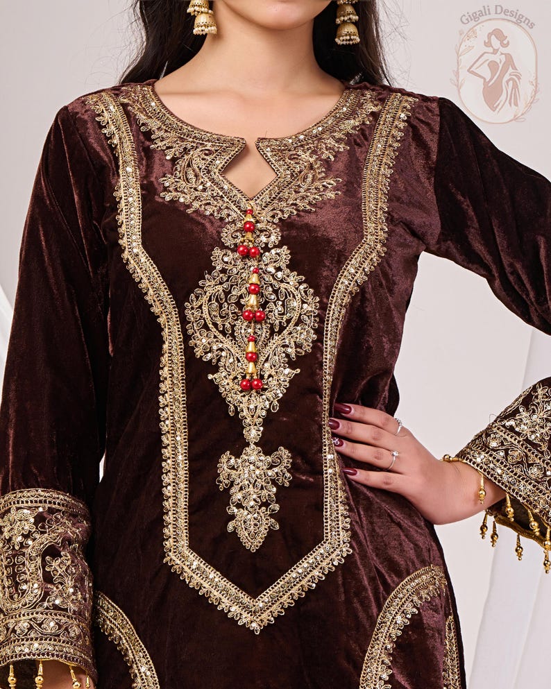 May include: A brown velvet tunic with intricate gold embroidery around the neckline, bodice, and cuffs. The design includes a keyhole neckline and is embellished with red beads. The sleeves are long, and the garment has a luxurious appearance.