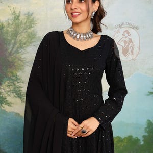 May include: A woman in a black, long-sleeved dress with a V-neck and detailed embroidery. She is wearing a matching black shawl, silver necklace, earrings, and rings. The background shows a painted landscape. The dress is from Gigali Designs.