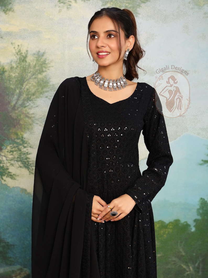 May include: A woman in a black, long-sleeved dress with a V-neck and detailed embroidery. She is wearing a matching black shawl, silver necklace, earrings, and rings. The background shows a painted landscape. The dress is from Gigali Designs.