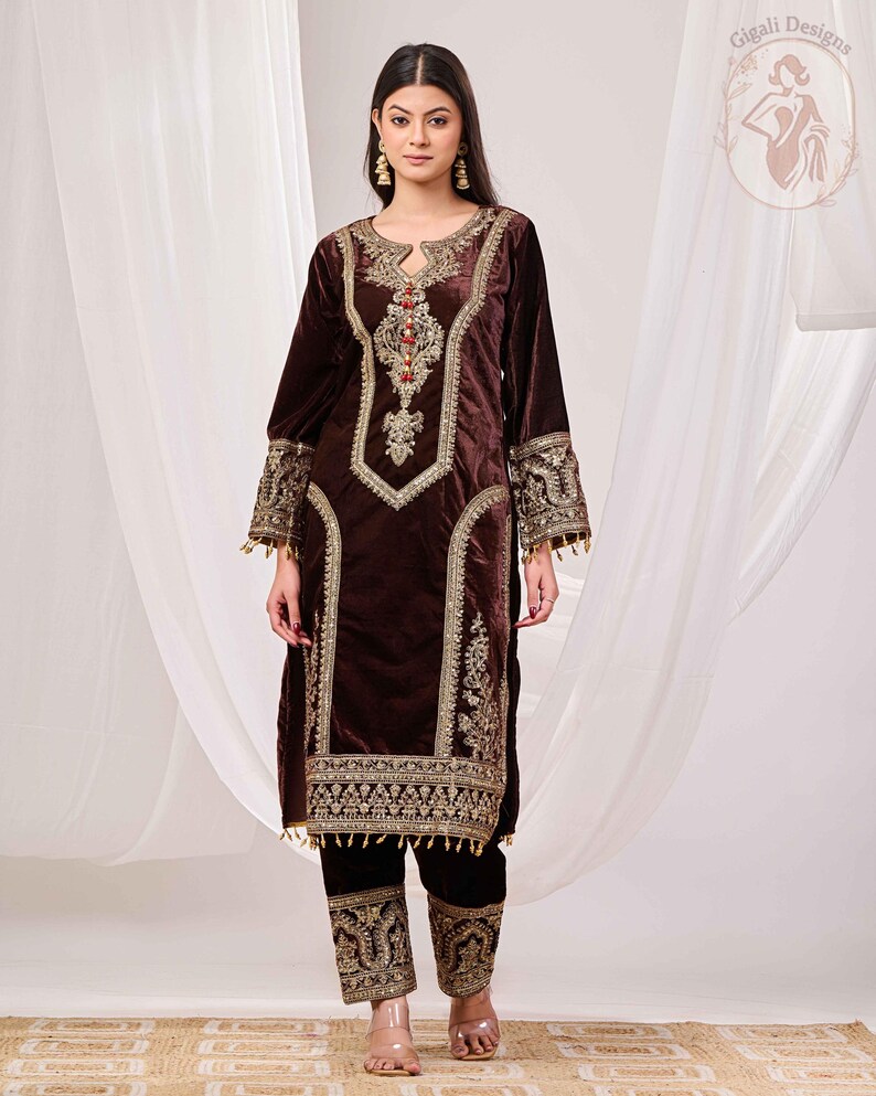 May include: A dark brown velvet outfit with intricate gold embroidery. The long-sleeved top features a keyhole neckline and ornate detailing around the collar, cuffs, and front panels. The matching pants have embroidered cuffs. The outfit is from Gigali Designs.