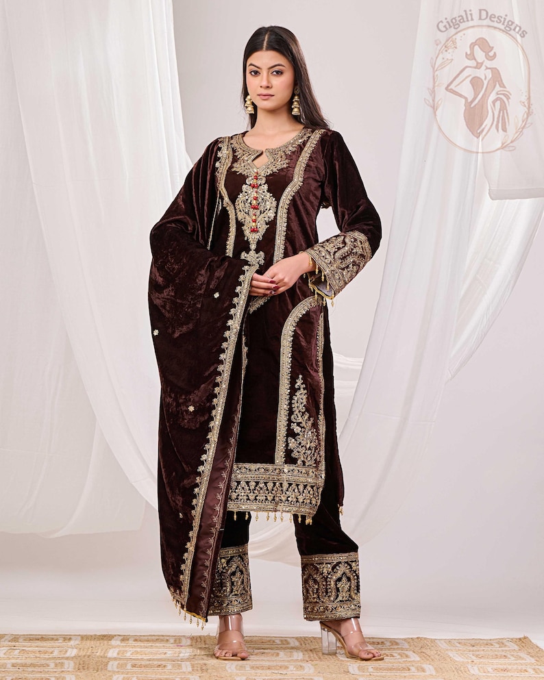 May include: A dark brown velvet salwar kameez with intricate gold embroidery. The ensemble includes a long-sleeved tunic, matching pants, and a shawl. The outfit features detailed embellishments around the neckline, cuffs, and hem.