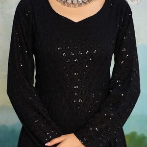 May include: A black, long-sleeved dress with a V-neck and detailed sequin and embroidered patterns. The dress has a fitted bodice and a flowing skirt. A silver necklace with decorative elements is worn around the neck.