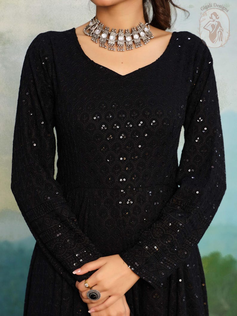 May include: A black, long-sleeved dress with a V-neck and detailed sequin and embroidered patterns. The dress has a fitted bodice and a flowing skirt. A silver necklace with decorative elements is worn around the neck.