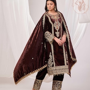 May include: A dark brown velvet salwar kameez set with intricate gold embroidery. The set includes a long-sleeved tunic, matching pants, and a flowing shawl. The outfit features detailed embellishments around the neckline, cuffs, and hem.