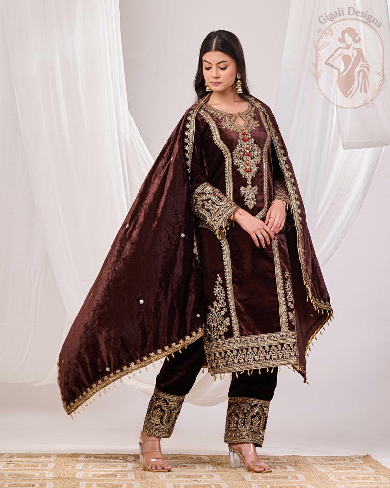 May include: A dark brown velvet salwar kameez set with intricate gold embroidery. The set includes a long-sleeved tunic, matching pants, and a flowing shawl. The outfit features detailed embellishments around the neckline, cuffs, and hem.