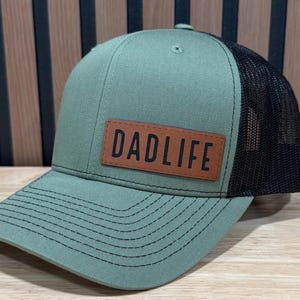 May include: Olive green and black trucker hat with a curved peak. The front panel has a brown leather patch with the text "DADLIFE". The hat has a mesh back and a snap closure.
