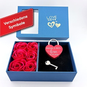 Op de afbeelding: Blauwe geschenkdoos met een hartvormig hangslot en sleutel, en zes rode roosvormige zeepjes. Het slot is gegraveerd met namen en een datum. Het deksel van de doos heeft het woord "LOVE" en hartmotieven.