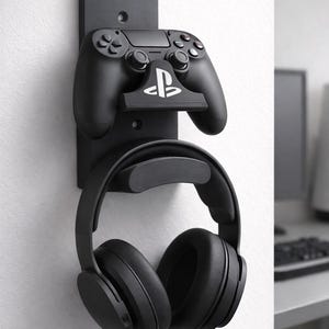Könnte beinhalten: Schwarzer, an der Wand montierter Gaming-Zubehörhalter. Der Halter verfügt über eine Controller-Halterung mit dem PlayStation-Logo und einen Kopfhörerhalter. Controller und Kopfhörer sind schwarz. Der Halter ist an einer weißen Wand montiert.