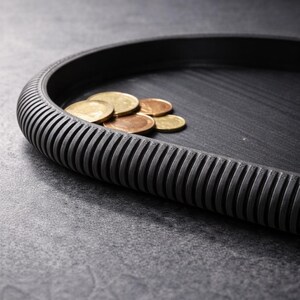 Peut inclure: Un plateau noir de forme ovale avec un bord côtelé et texturé. Plusieurs pièces de monnaie dorées et cuivrées sont à l'intérieur. Le plateau a une finition mate.