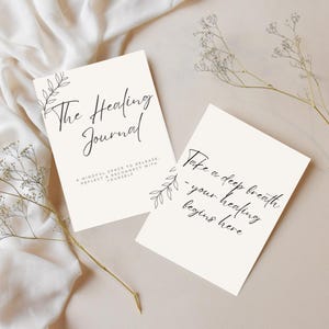 Könnte beinhalten: Zwei cremefarbene Journale mit schwarzer Schrift und botanischen Illustrationen. Auf einem Journal steht "The Healing Journal" und auf dem anderen "Take a deep breath - your healing begins here."