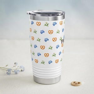 Könnte beinhalten: Ein weißer Edelstahlbecher mit einem wiederkehrenden Muster aus braunen Brezeln, blauen Vergissmeinnicht-Blüten und kleinen weißen Blumen. Der Trinkbecher verfügt über einen transparenten Kunststoffdeckel und einen silbernen Metallboden.