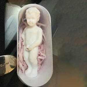 Puede incluir: Una figura blanca de un niño pequeño de pie, presentada en una caja blanca con relleno de papel rosa. La figura tiene un acabado liso y es de color crema claro. La caja tiene una parte superior curva.
