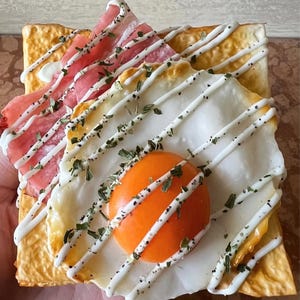 Op de afbeelding: Een vierkant geroosterd brood belegd met een gebakken ei, spek en een scheut witte saus. De eidooier is fel oranje en het spek is roze. Bestrooid met groene kruiden, is dit gerecht een visueel aantrekkelijk ontbijt of brunch.