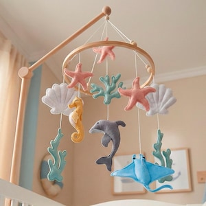 May include: A baby crib with a wooden mobile featuring felt sea creatures. The mobile includes a seahorse, dolphin, stingray, starfish, and coral in shades of blue, pink, yellow, and white. The crib is white, and a baby is sleeping under a blue blanket.