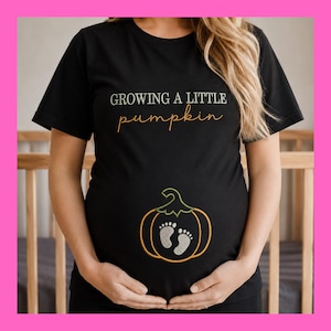 May include: A black short-sleeved t-shirt featuring the text GROWING A LITTLE pumpkin in white and orange lettering. Below the text is an embroidered graphic of an orange pumpkin outline containing a pair of white baby footprints inside.